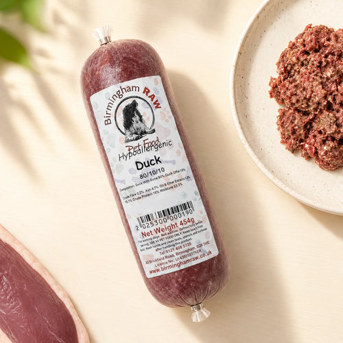 A tube of Birmingham Raw Duck raw dog food, shown on a light beige surface next to raw meat
