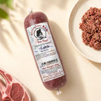 A tube of Birmingham Raw Lamb raw dog food, shown on a light beige surface next to raw meat