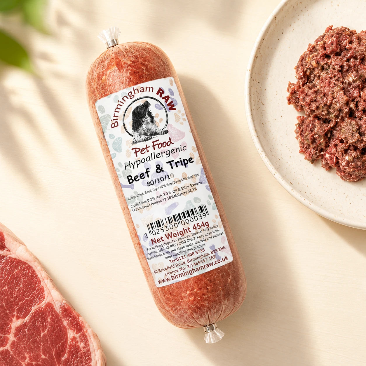 A tube of Birmingham Raw Beef And Tripe raw dog food, shown on a light beige surface next to raw meat