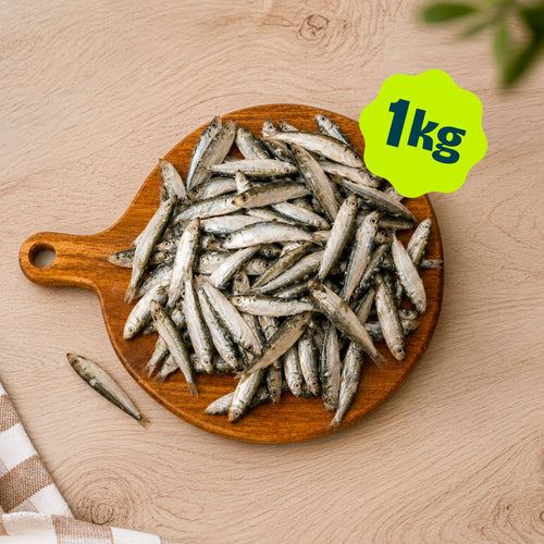 A pile of Birmingham Raw frozen sprat dog treats sold by the kilogram, displayed on a round wooden cutting board over a light wood kitchen unit.