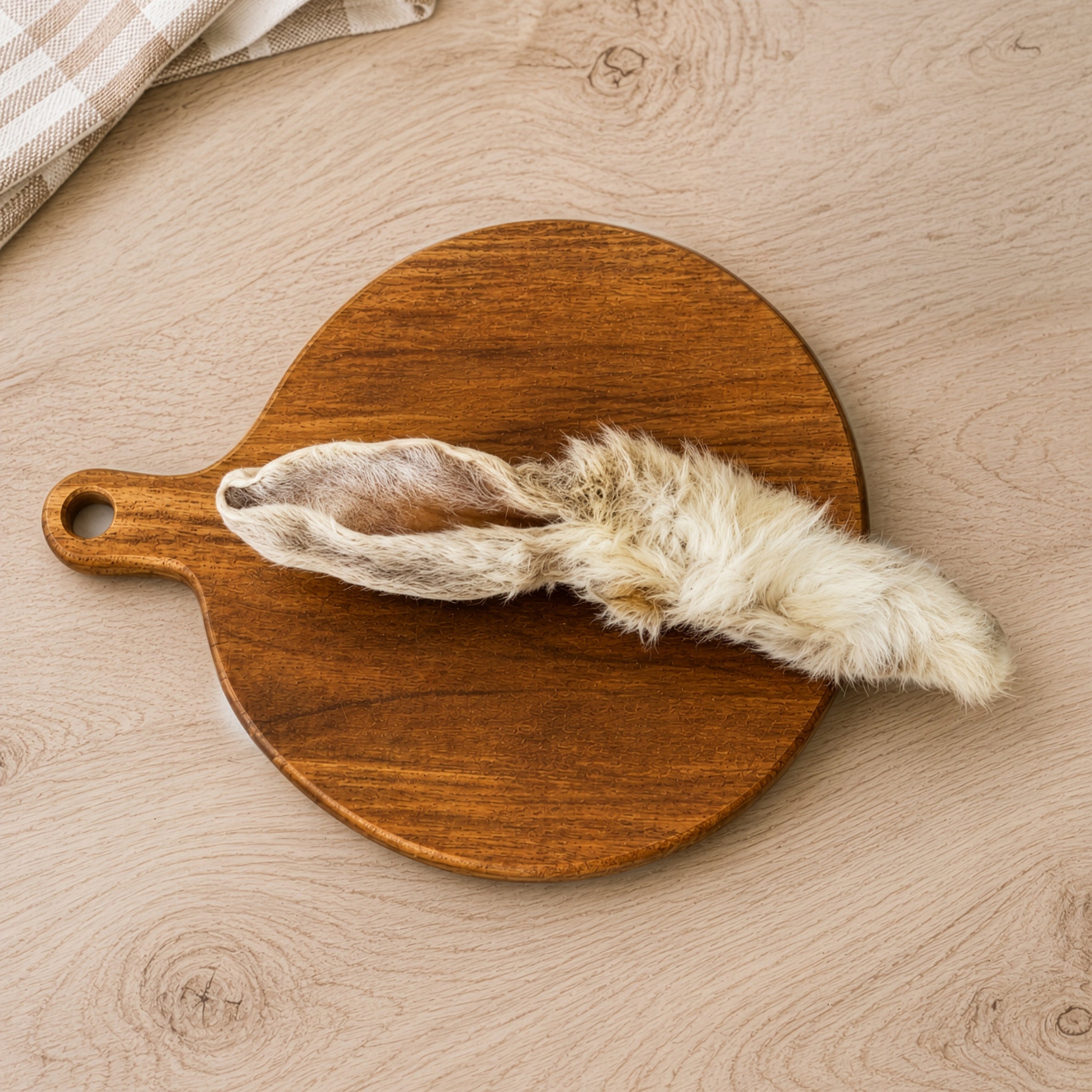 A hair rabbit ear dog treat, displayed on a round wooden cutting board over a light wood kitchen unit.