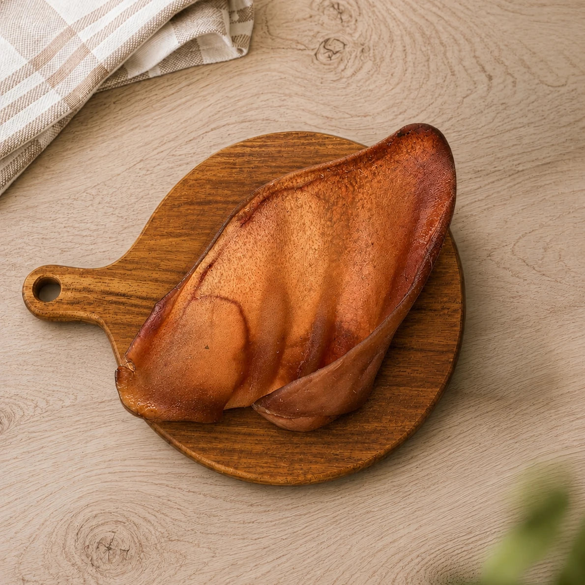 A dried pig ear dog treat, displayed on a round wooden cutting board over a light wood kitchen unit.